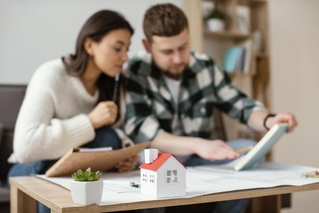 Casal jovem sentado à mesa de casa analisando documentos e projetos, com um pequeno modelo de casa em miniatura em destaque sobre a mesa.