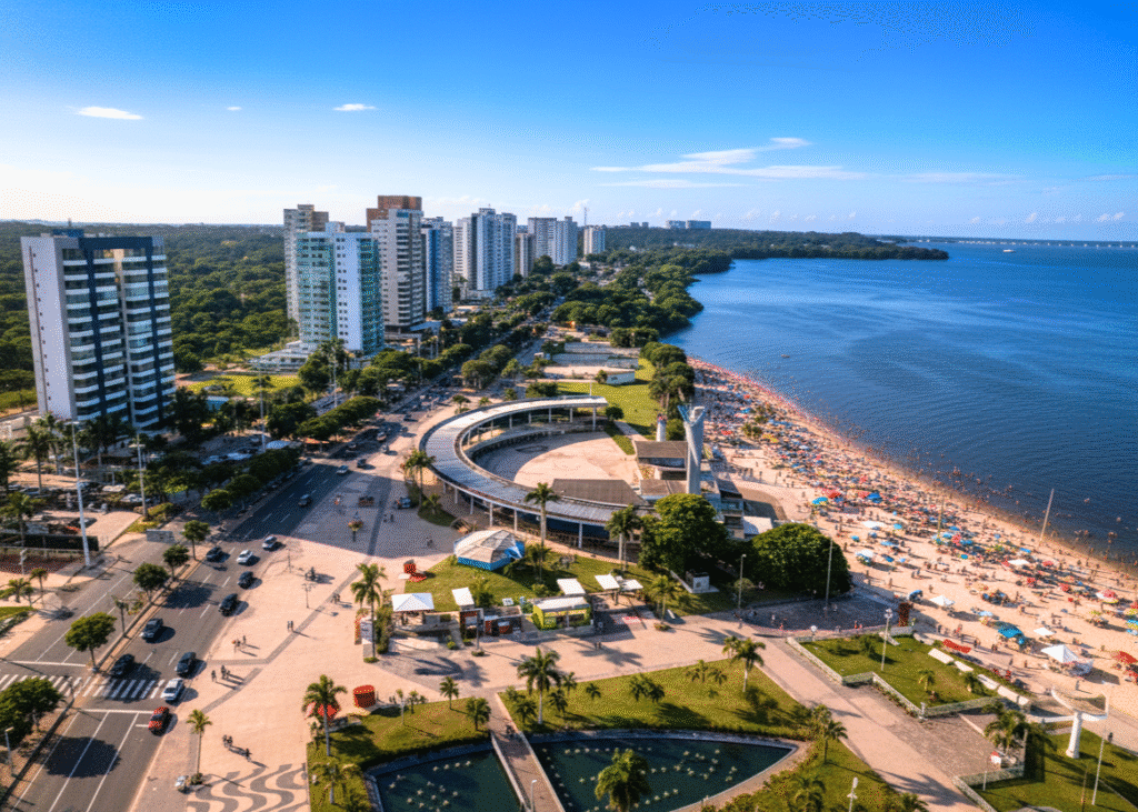 Vista aérea panorâmica da Praia da Ponta Negra em Manaus durante o dia, mostrando a extensa faixa de areia lotada de pessoas e guarda-sóis coloridos. À esquerda, uma avenida movimentada com carros e pedestres, ladeada por prédios residenciais altos e modernos. Ao centro, uma estrutura circular de lazer e áreas verdes com palmeiras. O vasto Rio Negro se estende ao longo da praia até o horizonte, sob um céu azul claro com poucas nuvens.