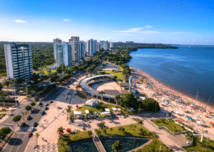 Vista aérea panorâmica da Praia da Ponta Negra em Manaus durante o dia, mostrando a extensa faixa de areia lotada de pessoas e guarda-sóis coloridos. À esquerda, uma avenida movimentada com carros e pedestres, ladeada por prédios residenciais altos e modernos. Ao centro, uma estrutura circular de lazer e áreas verdes com palmeiras. O vasto Rio Negro se estende ao longo da praia até o horizonte, sob um céu azul claro com poucas nuvens.