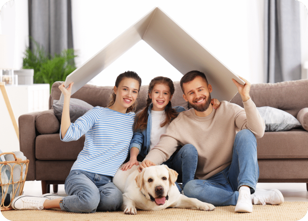 Foto de uma família feliz e sorridente (pai, mãe e filha pequena) sentada no chão da sala de estar com seu cachorro labrador deitado à frente. O casal segura um pedaço de papel ou papelão sobre as cabeças, formando um telhado simbólico, representando a ideia de um lar ou casa.