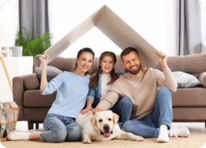 Foto de uma família feliz e sorridente (pai, mãe e filha pequena) sentada no chão da sala de estar com seu cachorro labrador deitado à frente. O casal segura um pedaço de papel ou papelão sobre as cabeças, formando um telhado simbólico, representando a ideia de um lar ou casa.