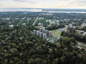 Vista aérea de um complexo residencial moderno composto por vários edifícios de altura média, integrado a uma vasta área de preservação ambiental da Floresta Amazônica. Ao fundo, vê-se a imensidão do Rio Negro e a linha do horizonte sob um céu de fim de tarde. A imagem destaca o equilíbrio entre o desenvolvimento urbano e a natureza preservada.