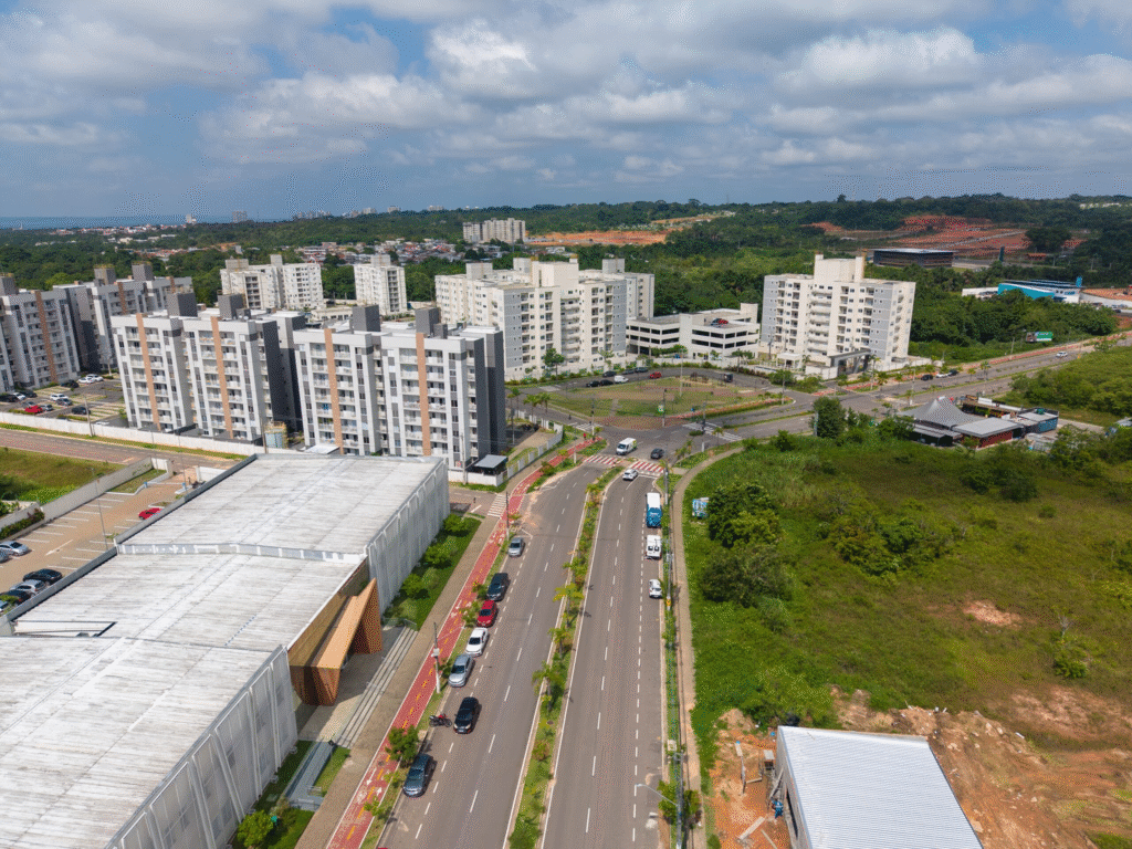 Vista aérea de uma avenida moderna em Manaus com pista dupla, sinalização atualizada e uma ciclovia pintada de vermelho à esquerda. Ao longo da via, observam-se diversos blocos de apartamentos residenciais de cor cinza e branco. O cenário inclui áreas de vegetação amazônica preservada ao fundo e terrenos em preparação para novas construções, sob um céu com nuvens brancas esparsas.