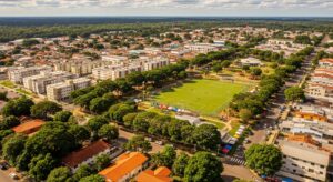 Vista aérea panorâmica de um bairro residencial arborizado e bem planejado. No centro da composição, destaca-se um campo de futebol gramado e áreas de lazer cercadas por densa vegetação. O entorno é composto por condomínios de prédios baixos e casas, com ruas organizadas. Ao fundo, uma vasta linha de horizonte com floresta preservada sob um céu azul com nuvens esparsas, transmitindo uma sensação de equilíbrio entre cidade e natureza.
